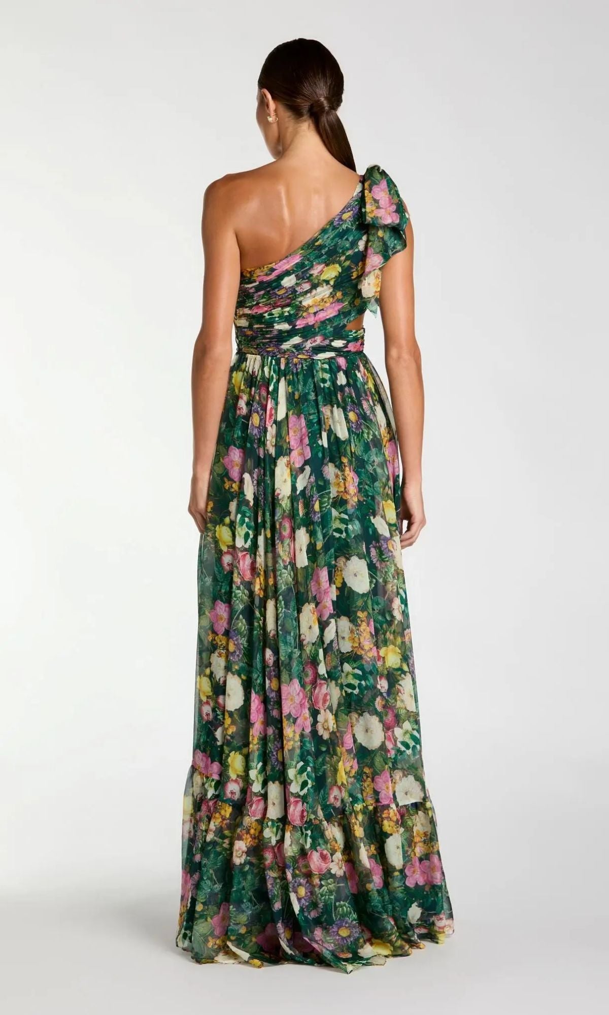 A woman faces away, wearing the Mac Duggal 68553 long formal dress—a one-shoulder evening gown featuring a colorful floral pattern on a green background.